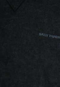 Tessuto grigio scuro con un sottile motivo testurizzato e piccolo testo bianco "DAILY PAPER" sul lato destro vicino a un collo a V.