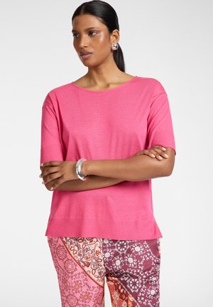 Vrouw met donker haar in een roze top met korte mouwen en meerkleurige gebloemde broek, draagt zilveren oorringen en een armband, armen gekruist.