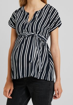 Zwangere vrouw draagt een zwart-wit gestreepte zwangerschapsblouse met verticale strepen en een gebonden riem, gecombineerd met een zwarte spijkerbroek.