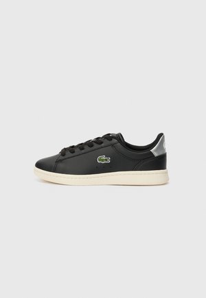 Baskets basses noires avec semelle blanche, lacets noirs, languette argentée au talon et petit logo crocodile vert sur le côté.