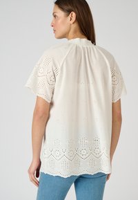 Blusa bianca con ricami a occhio di pernice, orlo scallop e maniche corte. Tessuto morbido con intricati motivi traforati.