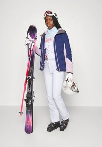 Equipamento de esqui com uma jaqueta isolada azul-marinho e rosa, camada base azul-claro, calças brancas, botas de esqui pretas, segurando esquis roxos e capacete branco.