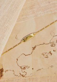 Bracciale d'oro con una catena sottile e una piastra centrale con un piccolo intaglio a forma di cuore, esposto su un tessuto testurizzato e uno sfondo chiaro.