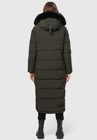 Langer olivgrüner Dämmermantel mit einem schwarzen Kunstfellkragen, gestepptem Design und einem Akzentgurt an der Rückseite. Getragen über schwarzen Hosen und Stiefeln.