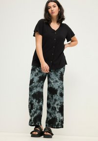 Černá krátká košile s knoflíky a véčkovým výstřihem, spárovaná s volnými kalhotami s batikovaným vzorem v černé a tyrkysové barvě, nošená s černými sandály.