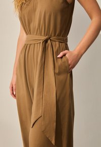 Pruun, varrukateta kombinesoon, millel on seotud vöökoht, küljetasku ja sile tekstuur. Kangas langeb õrnalt, rõhutades lõõgastunud istuvust.