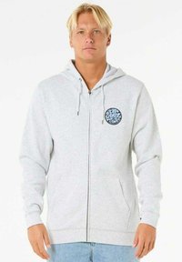 Haut à capuche gris zippé en tissu doux, doté d'une poche avant et d'un logo graphique circulaire sur la poitrine. Capuche ajustable avec cordon.