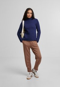 Marineblauer ribbed Strickkragenpullover, kombiniert mit hellbraunen tapered Pants und weißen Sneakern. Das Model steht vor einem schlichten Hintergrund.