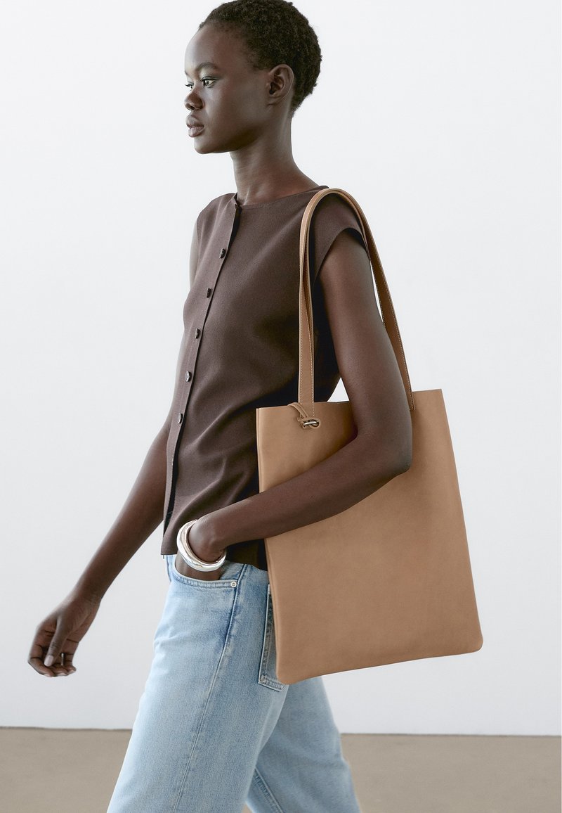 Tan leather tote bag with a minimalist design, smooth texture, and long handles, worn over a dark brown, button-up top with short sleeves.
