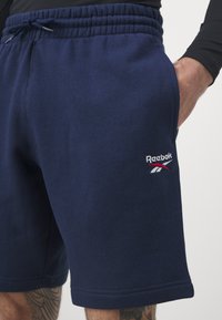 Tengerészkék Reebok rövidnadrág puha anyagból, rugalmas derékpánttal, oldalsó zsebből és fehér, piros hímzett logóval.