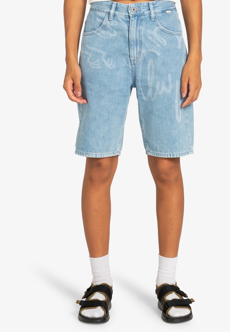 Personne portant un short en jean bleu clair avec des motifs graphiques pâles, des chaussettes blanches montantes et des sandales noires, debout sur fond blanc.