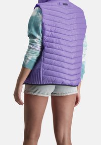 Fialová nadýchaná vesta s chevronovým prošíváním, lehký materiál, spárovaná s tričkem s dlouhým rukávem ve stylu tie-dye a denimovými šortkami.