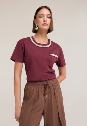 Femme portant un t-shirt bordeaux orné de strass au col et à la poche, associé à un pantalon marron taille haute à cordon, sur un fond uni.