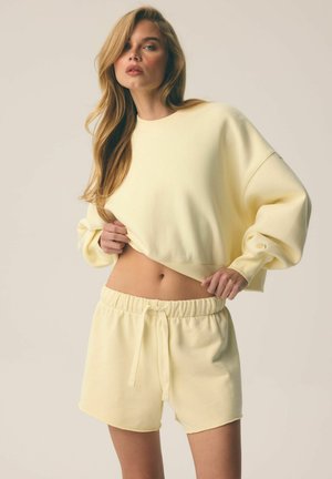 Kobieta z długimi blond włosami, ubrana w jasnożółtą, oversize'ową bluzę i dopasowane szorty ze sznurkiem, lekko podnosząca bluzę, aby odsłonić brzuch.