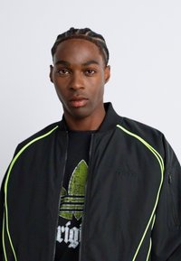 Ung mann med flettede hår som har på seg en svart Adidas-jakke med neon-gul piping over en svart skjorte med grønn Adidas-logo.