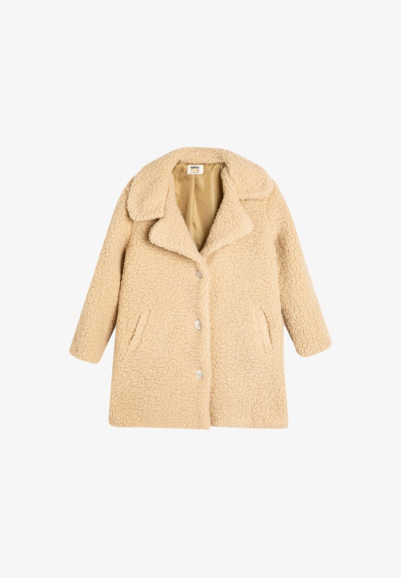 Beige tekoturkki, jossa on viistetty kaulus, napituksella ja sivutaskuilla. Teksturoitu materiaali lisää lämpöä ja pehmeää ilmettä.