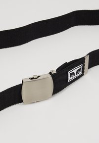Cinturón negro tejido con una hebilla de metal plateado mate. Presenta una forma rectangular y una etiqueta de tela con un detalle del logo de dos manos.