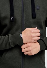 Felpa verde con zip, con lacci neri, polsini a costine e una patch con logo sul petto. Materiale morbido, vestibilità ergonomica e dettaglio della zip.