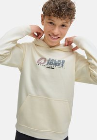 Un garçon aux cheveux bouclés sourit en ajustant la capuche d'un sweatshirt crème arborant le logo "Jack & Jones" sur le devant.