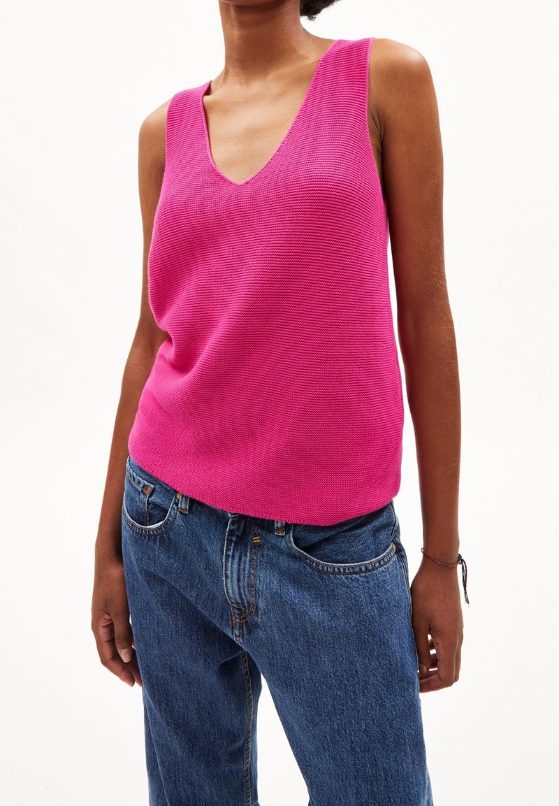 Kvinna i ärmlös ljusrosa stickad topp med V-ringning och blå jeans mot vit bakgrund.