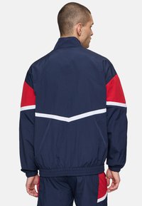 Giacca a vento navy con accenti rossi e bianchi. Presenta un colletto alto, polsini elasticizzati e un motivo a zigzag sulla schiena.