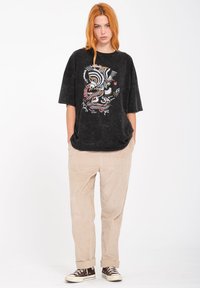 Camiseta negra de gran tamaño con un diseño gráfico colorido, combinada con pantalones de pana beige y zapatillas marrones. Un atuendo simple y casual.