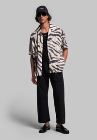 Camicia a maniche corte con bottoni, di base crema e strisce a zebra nere, abbinata a pantaloni neri a gamba larga e scarpe nere. Cappello nero e occhiali da sole.