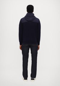 Marinblå stickad sweatshirt med kontrasterande huva, kombinerad med mörka jeans och vita sneakers. Bakifrån visas en figurnära design och ribbad mudd.