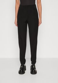 Pantalons noirs ajustés en tissu lisse, au design fuselé et avec des plis avant. Portés avec des chaussures à plateforme noires imposantes.