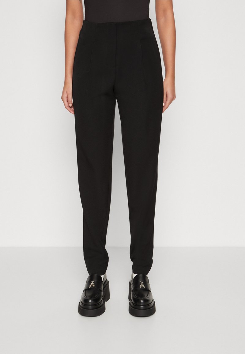 Pantalons noirs ajustés en tissu lisse, au design fuselé et avec des plis avant. Portés avec des chaussures à plateforme noires imposantes.