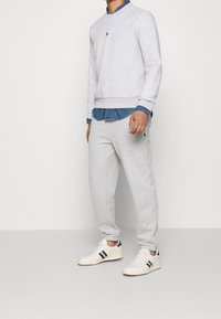Sudadera gris claro con cuello redondo, acompañada de pantalones de chándal gris claro. Zapatillas blancas con rayas verdes y negras. Camisa de mezclilla debajo.