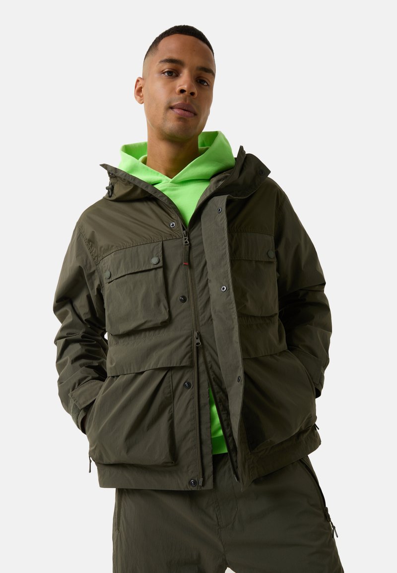 Olive green, waterproof jacket with a hood, featuring two front flap pockets, a zipper, and a drawstring hem over a neon green hoodie.