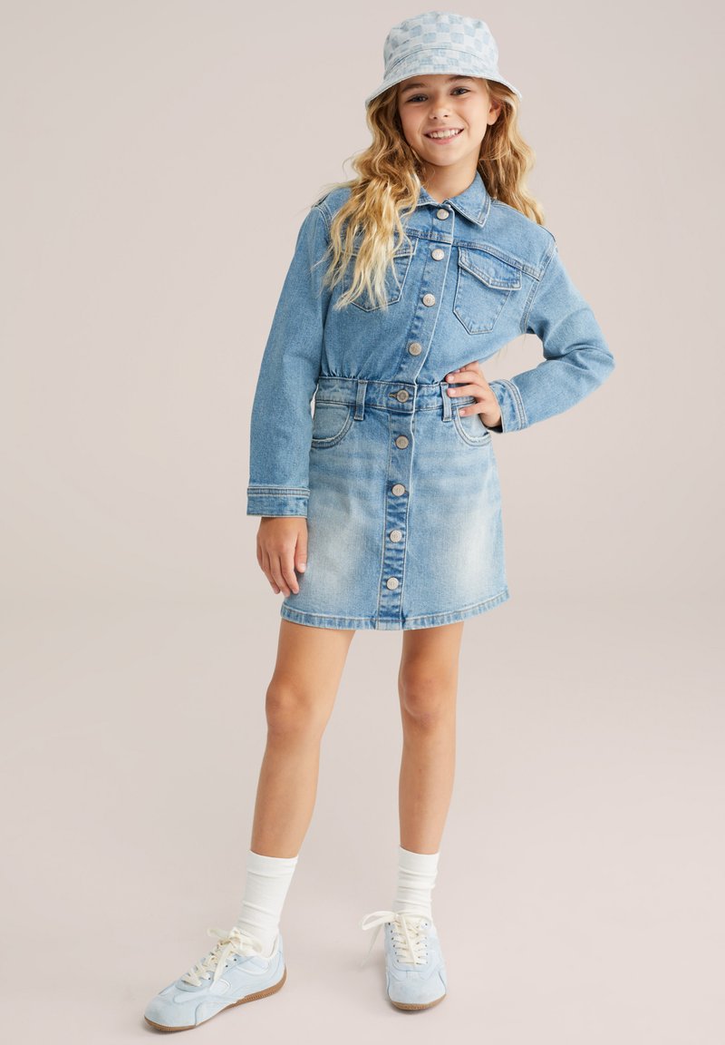 Meitene smaidot ar gariem viļņainiem matiem, ģērbta džinsa jaciņā, baltās zeķēs, gaismas zila apavos un pārbaudītu rakstu cepurē.