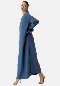 Długa, zwiewna niebieska sukienka z miękkiego materiału, z długimi rękawami ze zgromadzonymi mankietami i bocznymi rozcięciami, zestawiona z przezroczystymi sandałami na obcasie.