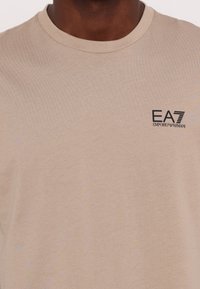 Béžové tričko s krátkým rukávem vyrobené z jemné bavlny. Má kulatý výstřih a malé černé logo EA7 Emporio Armani na levé hrudi.