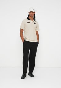 Beige polo shirt with a black collar, logo patches, paired with black trousers and black shoes; model wearing a white cap.