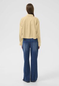 Skrojona, jasnobeżowa koszula z długim rękawem, z plisą z tyłu i spiczastym kołnierzykiem, zestawiona z jeansami z wysokim stanem i szerokimi nogawkami w kolorze niebieskim.