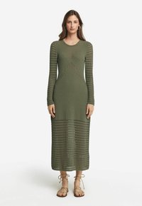 Stickad klänning med lång ärm i olivgrön med mönster av öppet arbete. Figurformad överdel, avslappnad kjol och knälång längd. Bärs med sandaler som knyts runt vristen.