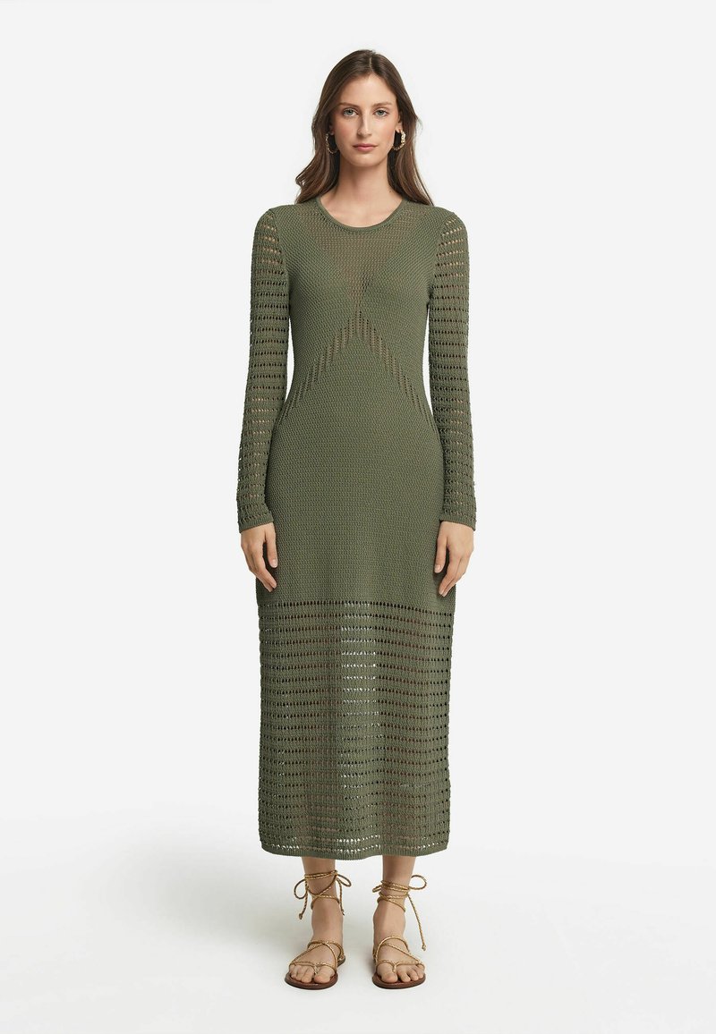 Stickad klänning med lång ärm i olivgrön med mönster av öppet arbete. Figurformad överdel, avslappnad kjol och knälång längd. Bärs med sandaler som knyts runt vristen.