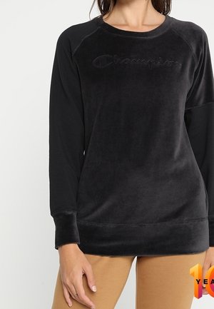 Kvinde iført en sort langærmet Champion sweatshirt og beige bukser, stående mod en ensfarvet baggrund.