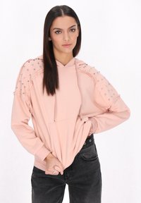 Hoodie rosa chiaro con tasca frontale, caratterizzato da borchie argentate sulle spalle. Ha una vestibilità comoda e cordini sul collo.