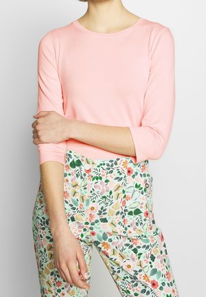 Femme portant un haut rose à manches longues et un pantalon taille haute à imprimé floral, debout avec un bras croisé sur son corps.