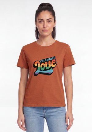Femme aux cheveux foncés attachés, portant un T-shirt couleur rouille avec un texte "Celebrate Love" aux couleurs de l'arc-en-ciel et un jean bleu clair, debout, immobile.