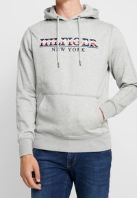 Uomo che indossa un felpa grigia chiara con la scritta "HILFIGER NEW YORK" e jeans blu, mano destra in tasca, mano sinistra rilassata al fianco.