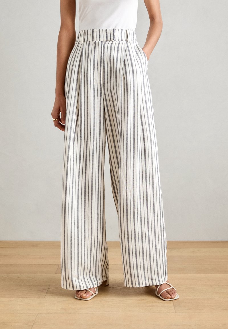 Femme portant un pantalon taille haute à jambes larges avec des rayures verticales noires et blanches, associé à des sandales blanches à brides et un haut blanc.