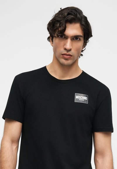Νέος άνδρας με σγουρά σκούρα μαλλιά που φοράει ένα μαύρο t-shirt Moschino Active, κοιτάζοντας κατευθείαν μπροστά σε ένα απλό φωτεινό φόντο.