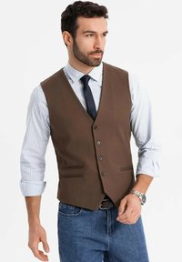 Mann trägt eine braune Knopf-Weste über einem hellblauen gestreiften Hemd mit hochgekrempelten Ärmeln, einer blauen Krawatte, blauen Jeans und einer silbernen Armbanduhr.