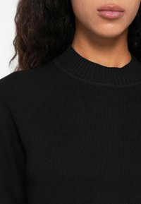 Donna con capelli ricci scuri che indossa un maglione a coste nero con scollo rotondo, inquadrata dalle spalle alle labbra.