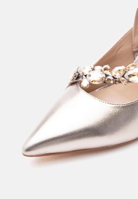 Μυτερό μεταλλικό χρυσό φλατ παπούτσι με διακοσμητικό λουράκι που διαθέτει διακοσμήσεις από διαφανείς και κεχριμπαρένιες πέτρες.