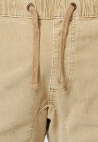 Les joggers en velours côtelé beige présentent une finition texturée, une taille élastique et des cordons de serrage coordonnés. Des coutures visibles ajoutent du détail.